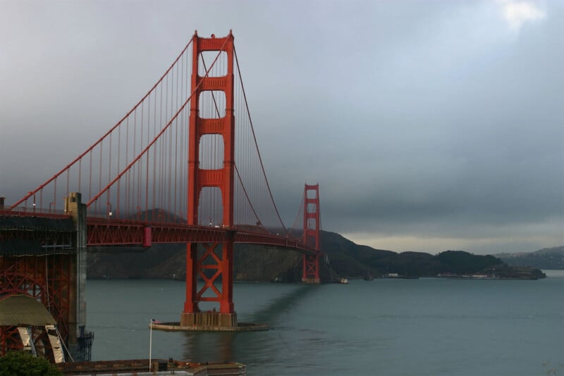 A view of the Golden Gate Bridge spanning the water with a cloudy sky.  The iconic red and orange towers and cables of the bridge contrast with the gloomy background.  Distant coasts and hills are faintly visible under the gray clouds.