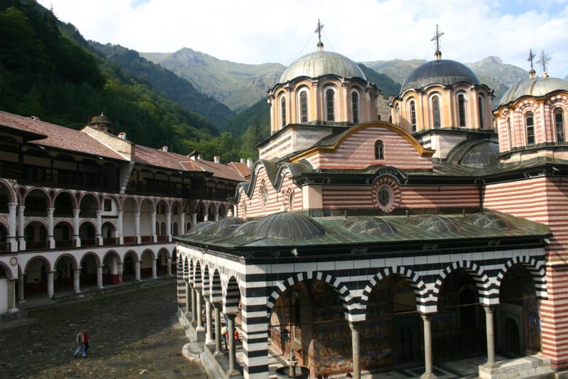 The image shows the Rila Monastery in Bulgaria, with its distinctive striped facade, domed roofs and courtyard.  The surrounding green mountains evoke a peaceful atmosphere.  Several people can be seen walking around the courtyard, showing off the monastery's grandeur and architectural details.